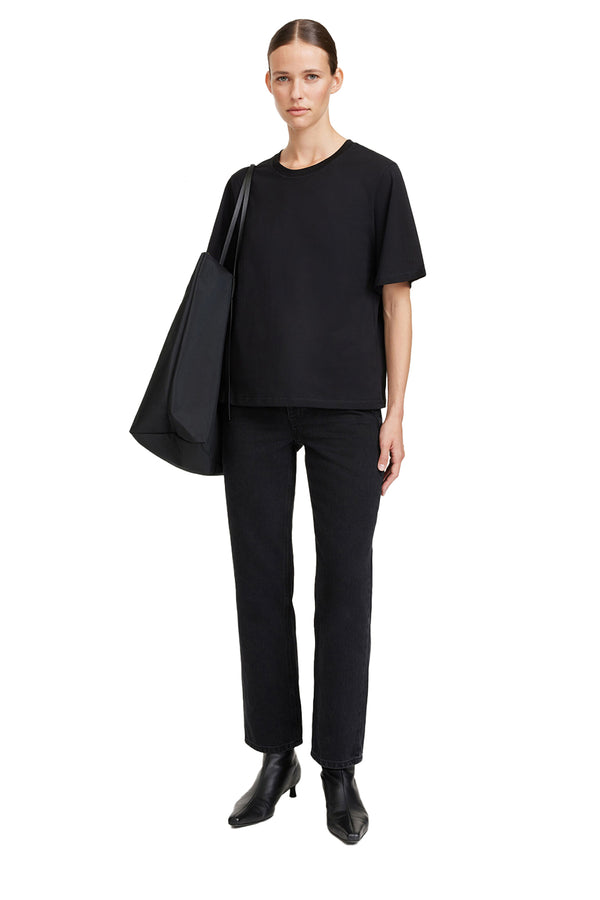 Front view of a woman wearing By Malene Birger t-shirt in black, paired with black trousers, black shoulderbag and a pair of black heels, shown against a plain white background.